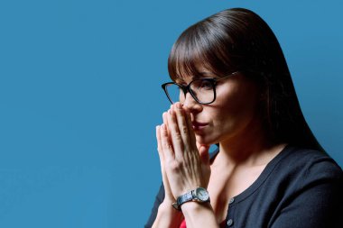 Üzgün, üzgün, gözlüklü, elleri katlanmış, mavi arka planı kopyalayan olgun bir kadının yakın çekimi. Yalnızlık sorunları, hastalık sorunları, depresyon, dua, akıl sağlığı, orta yaş