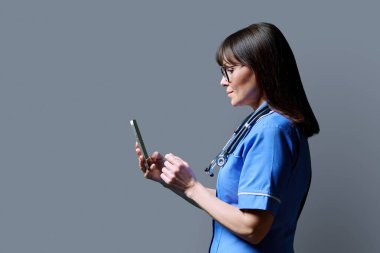 Gülümseyen kadın hemşire akıllı telefon kullanıyor, profil görüntüsü gri stüdyo arka planı, kopyalama alanı. Mobil uygulamalar, tıbbi hizmetlerdeki teknolojiler, sağlık, profesyonel yardım, tıbbi bakım