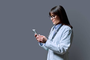 Beyaz laboratuvar önlüklü, gülümseyen, kendine güvenen kadın doktorun profil portresi. Gri stüdyo arka planında telefon akıllı telefonu kullanıyor. Kopya alanı. Sağlık, ilaç, personel, tedavi, tıbbi hizmetler, teknoloji
