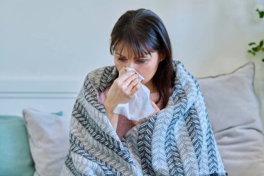 Hasta, olgun bir kadın. Burnunu sümkürerek hapşırıyor. Evdeki kanepede oturuyor. Soğuk algınlığı, sonbahar kışı mevsimsel solunum hastalıkları, tedavi ve bakım