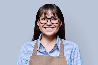 Gri arka planda önlüklü ciddi orta yaşlı kadın. Başarılı bir yetişkin kadın küçük işletme sahibi, kameraya bakan bir girişimci. İş adamları reklam işine başladı