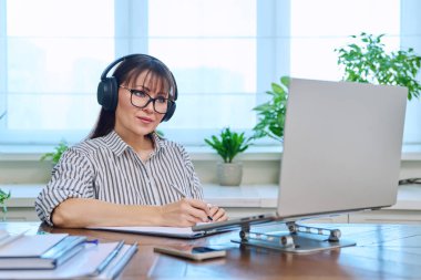 Merkez ofisteki bilgisayarda çalışan orta yaşlı kulaklıklı bir kadın web kamerasına bakarak konuşuyor. Çevrimiçi toplantı, uzak çalışma finansal danışmanlık danışmanlık blog yazarlığı, zihinsel terapi
