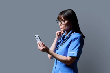 Akıllı telefon kullanan ciddi bir kadın hemşire. Gri stüdyo arka plan fotokopi alanının profil görüntüsü. Mobil uygulamalar tıbbi hizmetlerde teknolojiler, sağlık alanında profesyonel yardım tıbbi bakım kavramı