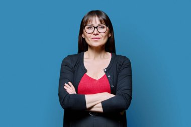 Mavi arka planda çapraz kollu, gülümseyen orta yaşlı bir kadının portresi. Gözlüklü kadın kameraya bakıyor. 40 'ların profesyonel, güzellik sağlık yaşam tarzı iş hizmetleri