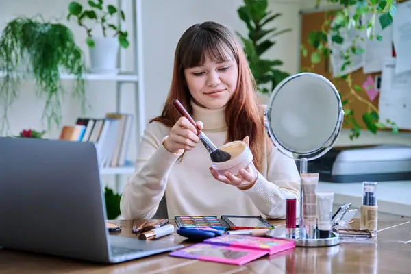 Young female showing cosmetics to laptop webcam, sitting at table at home. Beauty blog vlog, online course, training webinar on professional use of decorative cosmetics