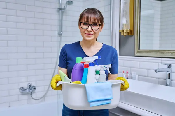 バスルームに洗剤の洗面台が付いている手袋の中年の女性 女性は 日常の家のクリーニング 職場でポーズをとるハウスクリーニングサービスワーカーのために準備します ハウスキーピングハウスワーククリーニングコンセプト — ストック写真