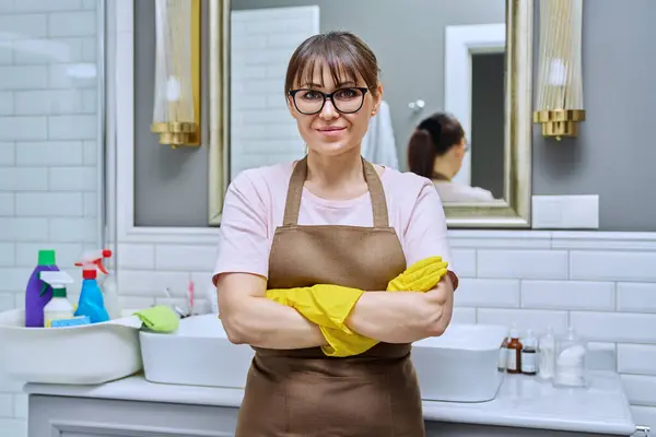 Middle-aged woman in gloves apron with crossed arms in bathroom. Female preparing for routine house cleaning, housecleaning service worker posing at workplace. Housekeeping housework cleaning