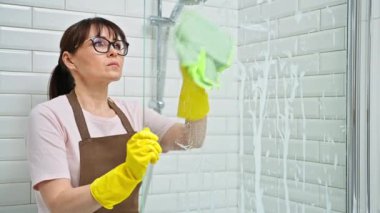 Önlüklü orta yaşlı bir kadın, profesyonel deterjanla banyo temizleyen eldivenler duşta cam temizliyor. Kadın ev hanımı temizlikçi, iş yerinde servis işçisi. Oda servisi ev işi
