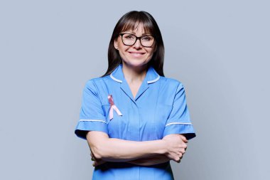 Mavi üniformalı gülümseyen kadın hemşire portresi gri arka planda Uluslararası Göğüs Kanseri Gününü sembolize eden pembe kurdeleli. Göğüs kanserinin tedavisi, tıp kadınlarının sağlık konsepti