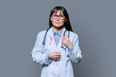 Gri arkaplanda Uluslararası Göğüs Kanseri Gününü sembolize eden pembe kurdeleli beyaz laboratuvar önlüklü gülümseyen kadın doktor portresi. Göğüs kanseri, ilaç, kadın sağlığı teşhisi