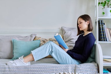 Evdeki kanepede rahatça oturup gazete okuyan genç bir kadın. Kurgu, hobi, eğlence, gençlik konsepti