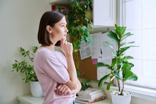 Serious young woman looking out the window, in home interior, thinking dreaming worrying female, profile view. Lifestyle, prayer, feelings, mental health, youth concept