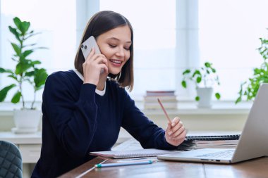 Mutlu gülümseyen genç bir kadın ofisteki masada bilgisayar laptopuyla oturuyor ve akıllı telefondan konuşuyor. Çalışan, uzaktan çalışan, serbest çalışan, öğretmen, öğrenci.