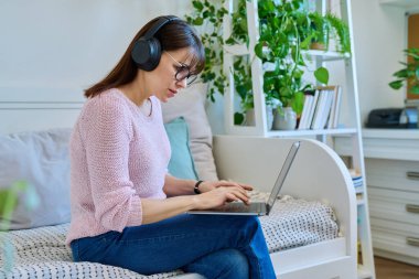 Kulaklıklı, dizüstü bilgisayarında yazı yazan, evdeki koltukta oturan orta yaşlı ciddi bir kadın. Boş zaman, iş, serbest çalışma, iletişim, eğlence teknolojileri.