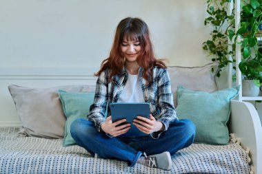 Genç, trend kız üniversite öğrencisi dijital tablet kullanarak serbest iletişim eğitimi alıyor, evdeki kanepede oturuyor. Teknoloji, gençlik, eğitim, yaşam tarzı kavramı