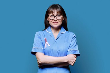 Mavi üniformalı gülümseyen kadın hemşire portresi mavi arka planda Uluslararası Göğüs Kanseri Gününü sembolize eden pembe kurdeleli. Göğüs kanserinin tedavisi, tıp kadınlarının sağlık konsepti