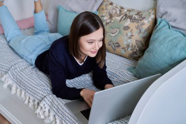 Dizüstü bilgisayar kullanan, klavyede yazan, evdeki kanepede yatan genç bir kadın. İş iletişimi eğitimi için internet çevrimiçi teknolojileri