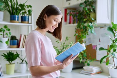 Mutlu gülümseyen, odaklanmış, pencerenin yanında kitap okuyan genç bir kadın. Önemli eğitim edebiyatı, ilginç kurgu, hobi kitapları.