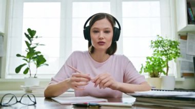 20 'li yaşlarda, konuşan, kulaklık takan, evde oturmuş kameraya bakan genç bir kızın webcam portresi. İnternet çevrimiçi webinar, görüntülü sohbet çağrı konferansı, teknoloji eğitimi