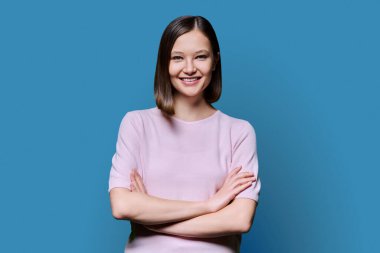 Mavi stüdyo arka planında çapraz kollu, kendine güvenen güzel bir kadın. 20 'li yaşlarda çekici bir kadın kameraya bakıyor. Yaşam tarzı, moda güzellik tarzı, gençlik iş eğitimi konsepti