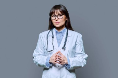 Steteskoplu beyaz laboratuar önlüklü ciddi orta yaşlı kadın doktor gri stüdyo geçmişindeki kameraya bakıyor. Sağlık, ilaç, personel, tedavi, tıbbi hizmetler kavramı