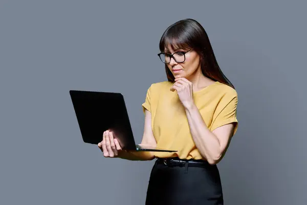 Middle-aged business serious woman using laptop on gray background. Mature successful female teacher mentor manager worker employee director. Internet online technologies, work, electronic teaching