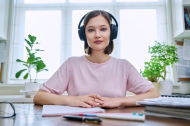 20 'li yaşlarda, kulaklık takan, evde oturmuş kameraya bakan ve dinleyen genç bir kızın webcam portresi. İnternet çevrimiçi webinar, görüntülü sohbet çağrı konferansı, teknoloji eğitimi
