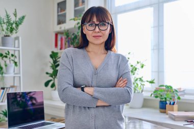 Gözlüklü, kendinden emin orta yaşlı bir kadının portresi. Kolları çapraz giyinmiş, iç mekândaki kameraya bakıyor. 40 'lı yaşlar, yaşam tarzı, sağlık, olgun kadın konsepti