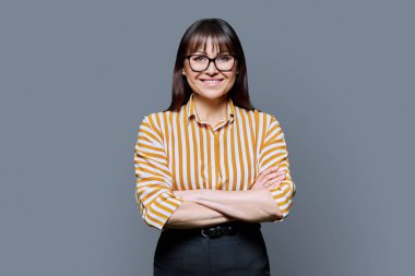 Gri arka planda çapraz kollu, güler yüzlü orta yaşlı iş kadınının portresi. Gözlüklü kadın kameraya bakıyor. 40 'ların profesyonel, güzellik sağlık yaşam tarzı iş hizmetleri