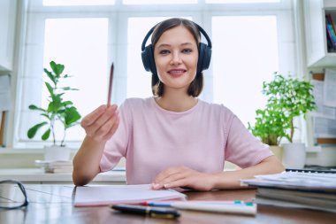 20 'li yaşlarda, kulaklık takan, masasında oturmuş kameraya bakıp konuşan genç bir kızın webcam portresi. İnternet çevrimiçi webinar, görüntülü sohbet çağrı konferansı, teknoloji eğitimi