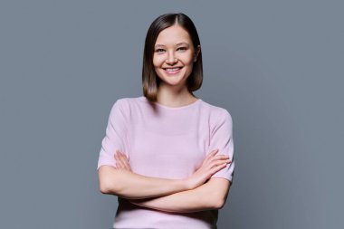 Gri stüdyo arka planında çapraz kollu güzel, kendine güvenen bir kadın. 20 'li yaşlarda çekici bir kadın kameraya bakıyor. Yaşam tarzı, moda güzellik tarzı, gençlik iş eğitimi konsepti