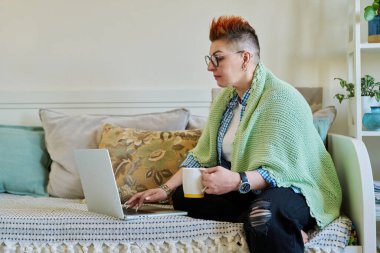 Sıcak battaniyeli sıcak bir içecek taşıyan olgun bir kadın evdeki kanepede oturan dizüstü bilgisayara bakıyor. Ev konforu, soğuk mevsimler, yaşam tarzı, iş iletişimi teknolojisi