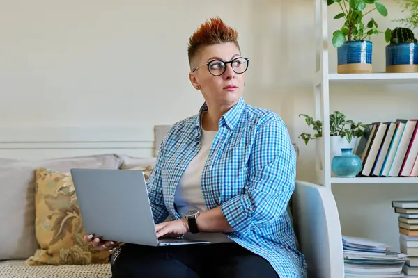 ラップトップを使用して自宅でソファーに座っている中年の女性 コンピュータで作業するカジュアルな服で赤い髪の真剣な女性 リラックス コミュニケーション — ストック写真