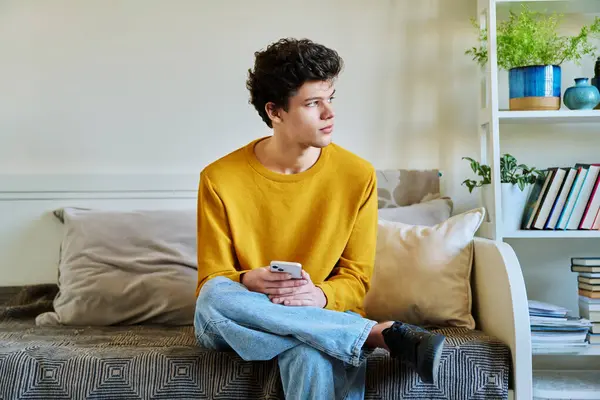 stock image Young guy holding smartphone in hands sitting on couch at home. Handsome university college student 19-20 years old in yellow with curly hair. Technologies, mobile apps, study communication leisure