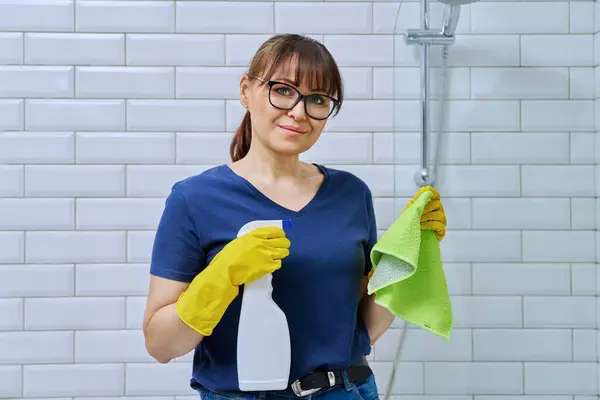 Middle-aged woman in gloves with detergent spray washcloth doing cleaning in bathroom, smiling looking at camera. Routine house cleaning, home hygiene, housecleaning service, housekeeping, housework