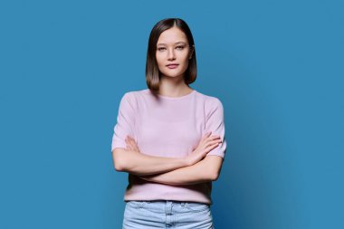 Mavi stüdyo arka planında çapraz kollu, kendine güvenen güzel bir kadın. 20 'li yaşlarda çekici bir kadın kameraya bakıyor. Yaşam tarzı, moda güzellik tarzı, gençlik iş eğitimi konsepti