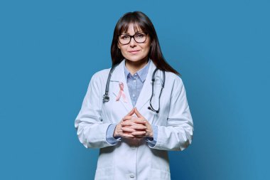 Mavi arka planda Uluslararası Göğüs Kanseri Gününü sembolize eden pembe kurdeleli beyaz önlüklü gülümseyen kadın doktor portresi. Göğüs kanseri, ilaç, kadın sağlığı teşhisi