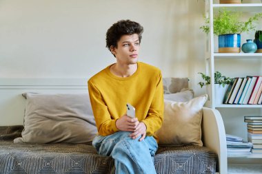 Evdeki koltukta oturan akıllı telefonu tutan genç adam. Yakışıklı üniversite öğrencisi 19-20 yaşlarında, sarı saçlı, kıvırcık saçlı. Teknolojiler, mobil uygulamalar, iletişim eğitimi