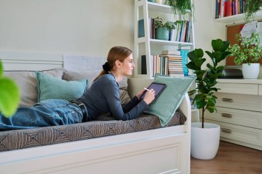 Dijital tabletli genç bir bayan, Stylus evde kanepede yatarken resim çiziyor. Teknolojiler, eğlence, yaratıcılık, eğitim, serbest çalışma, yaşam tarzı, gençlik kavramı