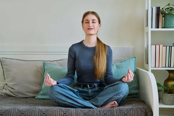 Young teenage calm relaxed female sitting in lotus position, with eyes closed meditating on couch at home. Health, mental and physical, lifestyle, youth concept