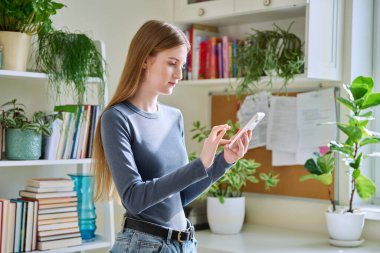 Akıllı telefon kullanan mutlu, gülümseyen genç kız, evde dikiliyor, 16,17, 18 yaşındaki kız mesajları okuyor. İletişim, eğlence, öğrenme ve alışveriş için modern dijital teknolojiler