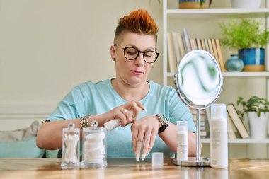 Evinde oturan aynalı orta yaşlı bir kadın. Yaşlanma önleyici cilt bakım ürünleri. Nemlendirici gıdaları temizlemek için. Güzellik çağı sağlık temizliği, 40 'lı yaşların ciltleri için tıbbi kozmetik ürünler.