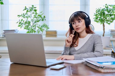 Kulaklıklı genç bir üniversite öğrencisi video konferansında sohbet ediyor bilgisayar ekranında ders veriyor, evdeki masada oturuyor. E-öğrenme, eğitim, teknoloji, bilgi