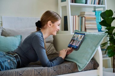 Genç bir kız öğrenci grup video konferansı yapıyor, dijital tablet kullanıyor, evdeki kanepede yatıyor. Kablosuz eğitim teknolojisi e-öğrenme sanal dersler uzaktan eğitim bilgileri