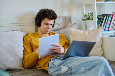 Kulaklıklı genç bir üniversite öğrencisi dizüstü bilgisayar kullanıyor, not alıyor, evdeki kanepede oturuyor. Webinar dinleme ses çevrimiçi konferans sohbet dersi e-öğrenme eğitim uzak sınıflar İnternet toplantıları