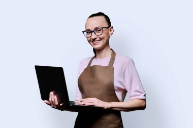 30 'lu yaşların portresi önlüklü mutlu bir kadın dizüstü bilgisayar kullanıyor, mavi stüdyo arkaplanındaki kameraya bakıyor. İşçi, girişim, küçük işletme, iş, hizmet sektörü, personel, gençlik kavramı