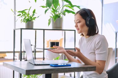Kulaklıklı genç bir kadın, kafede oturan dizüstü bilgisayarla görüntülü sohbet görüşmesi yapıyor. İnternet 'te okuyan kız üniversite öğrencisi Webinar dinlemek hazırlık sınavı, uzaktan çalışma