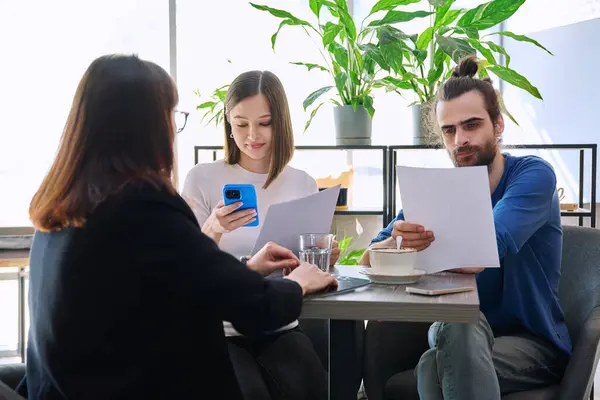 Stock image Couple of young clients at meeting with agent broker insurer realtor seller reading paper contract. Family customers tenants financing investment insurance rental offer, conclusion of purchase sale