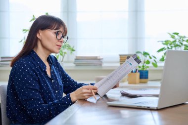 Orta yaşlı, ders kitabı olan bir kadın öğretmen ev ofisinde dizüstü bilgisayarla çalışıyor. Video konferansı uzaktan görüşme danışmanlığı ve eğitim teknolojisi kavramı