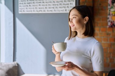 Genç, güzel, mutlu bir kadın elinde çay fincanıyla pencereden bakan fincan tabağıyla kafeterya kafeteryasının gri duvarını mesaj için fotokopi odası olarak kullanıyor. Kahve işi hizmetleri gençlik yaşam tarzı eğlencesi